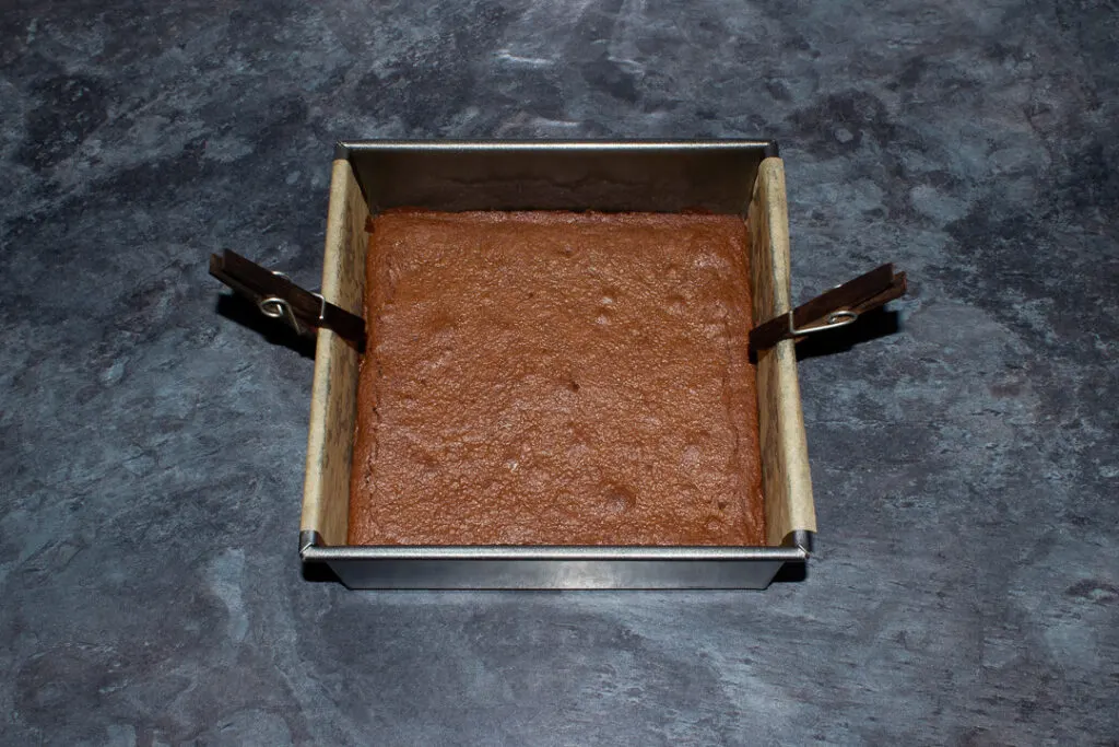 Baked brownie in a lined square baking tin on a kitchen worktop.