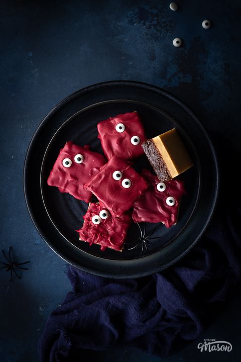 Flat lay close up of a dark blue plate filled with Halloween millionaire brownies set on a deep blue hand painted backdrop. There are also edible eyes, fake spiders and a blue linen napkin the background.