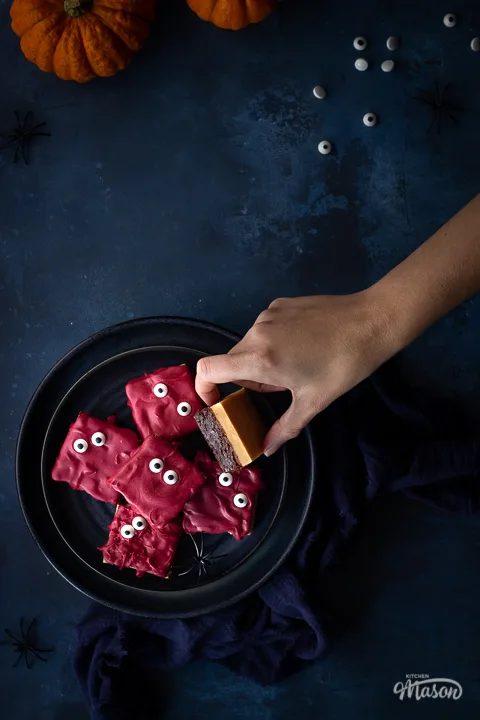 Flat lay view of a hand reaching in to a dark blue plate filled with Halloween millionaire brownies set on a deep blue hand painted backdrop. There are also two small pumpkins, a blue linen napkin edible eyes and fake spiders in the background.
