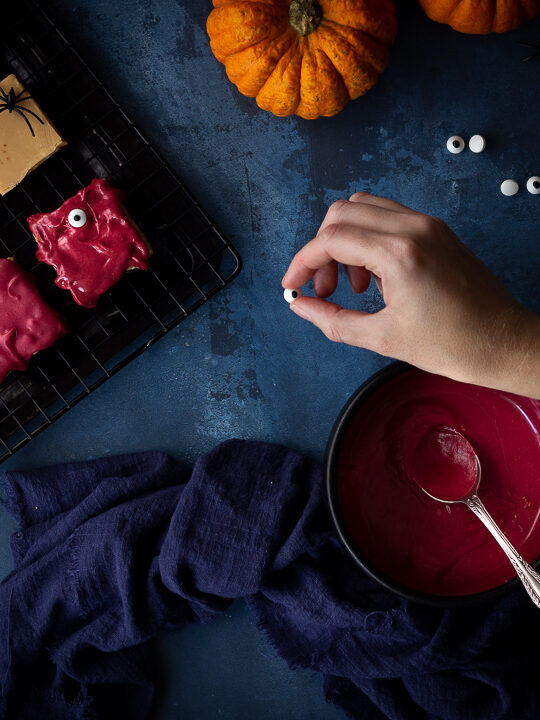 Flat lay view of Halloween millionaire brownies on a cooling rack being decorated with edible eyes. There is also a bowl of red white chocolate in a black bowl, a blue linen napkin, 2 small pumpkins and some edible eyes in the background. Set on a deep blue hand painted backdrop.