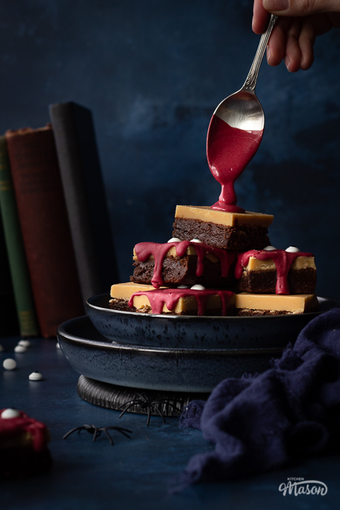 Front view of someone pouring melted red chocolate on a pyramid of Halloween Millionaire brownies, set on stacked blue plates against a deep blue hand painted backdrop. There is also a blue linen napkin, fake spiders, edible eyes and some old books in the background.