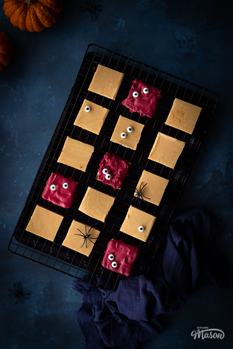 Flat lay view of Halloween millionaire brownies on a cooling rack set over a deep blue hand painted backdrop. There is also a blue linen napkin, two small pumpkins and fake spiders in the background.