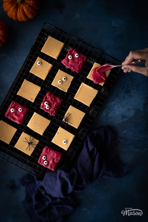 Flat lay view of Halloween millionaire brownies on a cooling rack being decorated with red white chocolate, set over a deep blue hand painted backdrop. There is also a blue linen napkin, two small pumpkins and fake spiders in the background.