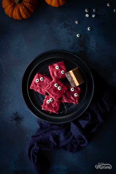 A dark blue plate filled with Halloween millionaire brownies set on a deep blue hand painted backdrop. There are also two small pumpkins, a blue linen napkin edible eyes and fake spiders in the background.