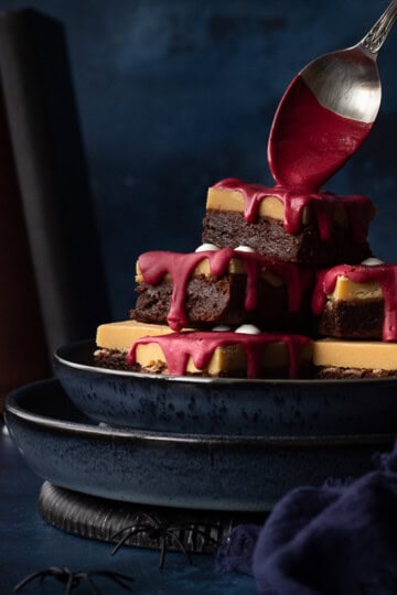 Front view of someone pouring melted red chocolate on a pyramid of Halloween Millionaire brownies, set on stacked blue plates against a deep blue hand painted backdrop. There is also a blue linen napkin, fake spiders, edible eyes and some old books in the background.