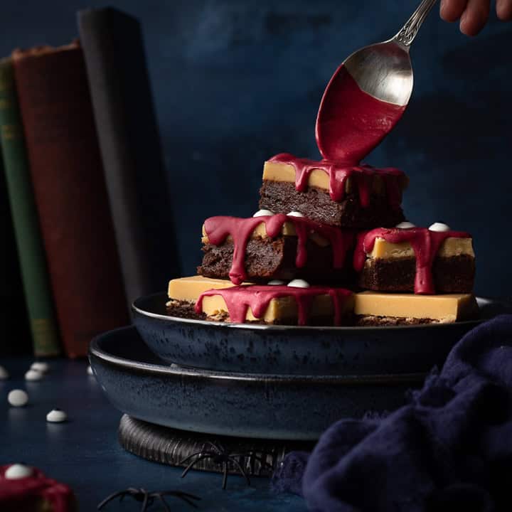 Front view of someone pouring melted red chocolate on a pyramid of Halloween Millionaire brownies, set on stacked blue plates against a deep blue hand painted backdrop. There is also a blue linen napkin, fake spiders, edible eyes and some old books in the background.