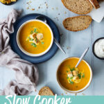 3 bowls of slow cooker carrot and coriander soup, one of which set on a blue plate, all topped with croutons, soured cream and coriander. There's a sliced loaf of bread, pot of soured cream, pot of croutons, spoons and a light brown linen napkin in the background. Set on a cool grey wood effect backdrop. A text overlay says "slow cooker carrot and coriander soup".