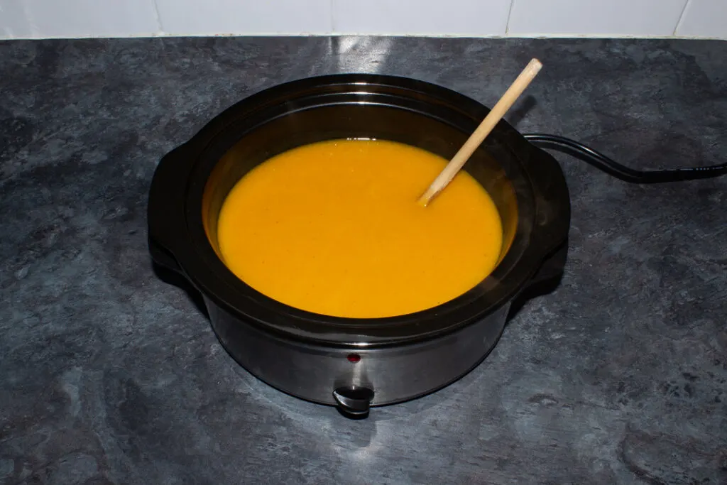 Cooked and blended carrot soup in a slow cooker on a kitchen worktop with a wooden spoon in it.