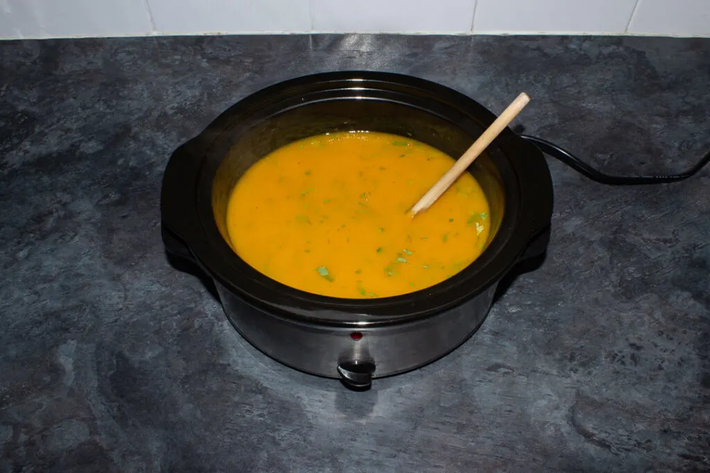 Cooked and blended carrot and coriander soup in a slow cooker on a kitchen worktop with a wooden spoon in it.