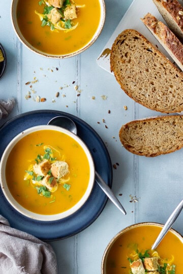 3 bowls of slow cooker carrot and coriander soup, one of which set on a blue plate, all topped with croutons, soured cream and coriander. There's a sliced loaf of bread, pot of soured cream, pot of croutons, spoons and a light brown linen napkin in the background. Set on a cool grey wood effect backdrop.