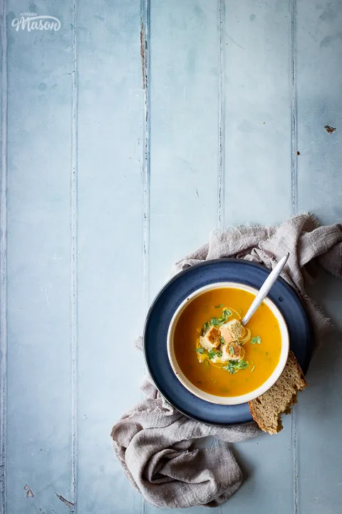 A bowl set on a blue plate over a light brown linen napkin, filled with carrot and coriander soup that's topped with croutons, soured cream and coriander. Set on a cool grey wood effect backdrop, there's also a spoon in the soup and a torn piece of bread on the side.