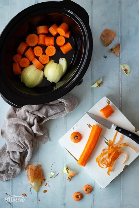A carrot being chopped on a wooden chopping board set on a cool grey wood effect backdrop. There's also a light brown linen napkin, a slow cooker filled with vegetables and some vegetable peelings in the background.