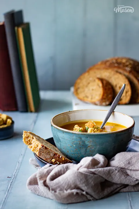 A front view of a bowl of slow cooker carrot and coriander soup with a spoon inside, topped with croutons, soured cream and coriander. Set on a light brown linen napkin over a cool grey wood effect backdrop, there's also a sliced loaf of bread, a pot of croutsons and some books in the background.