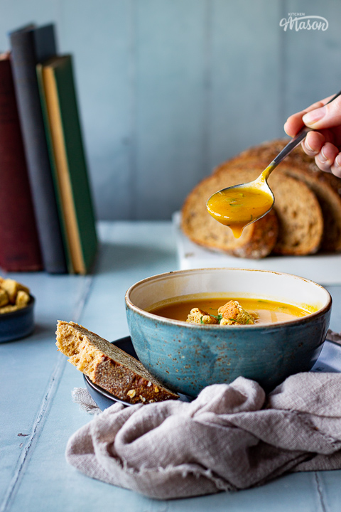 A front view of a spoon filled with soup over a bowl of slow cooker carrot and coriander soup, topped with croutons, soured cream and coriander. Set on a light brown linen napkin over a cool grey wood effect backdrop, there's also a sliced loaf of bread, a pot of croutsons and some books in the background.