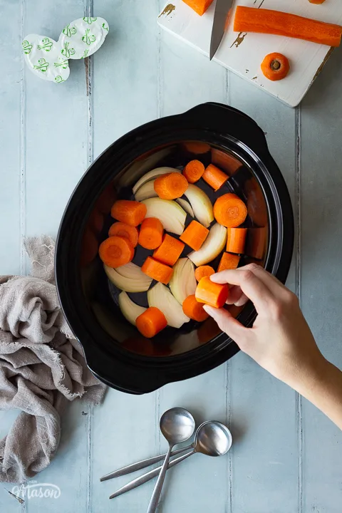 A hand placing a piece of carrot in a slow cooker that's filled with carrots and onions. There's a white chopping board with carrot on it, some stock pots, spoons and a light brown linen napkin in the background. Set on a cool grey wood effect backdrop.
