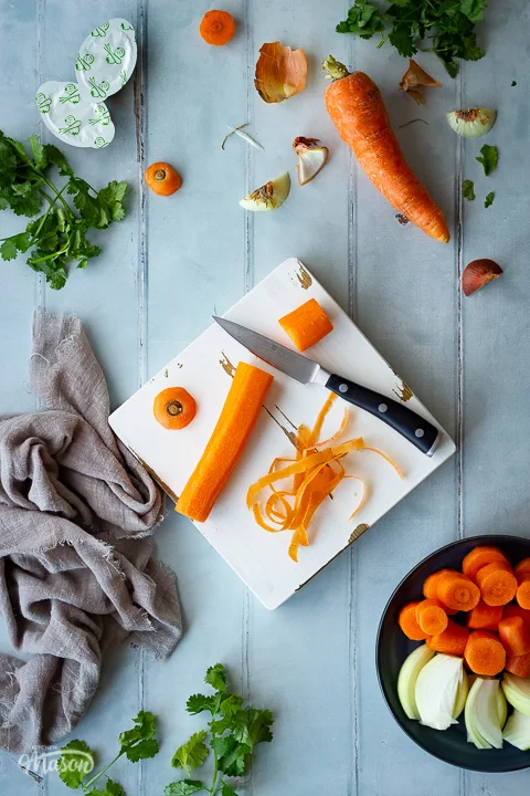 A chopped carrot on a white chopping board with a pairing knife. There's lots of vegetable peelings and coriander scattered around, a light brown linen napkin, some stock pots and a plate filled with chopped carrot and onion. All set on a cool grey wood effect backdrop.