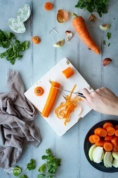 A carrot being chopped on a white chopping board with a pairing knife. There's lots of vegetable peelings and coriander scattered around, a light brown linen napkin, some stock pots and a plate filled with chopped carrot and onion. All set on a cool grey wood effect backdrop.
