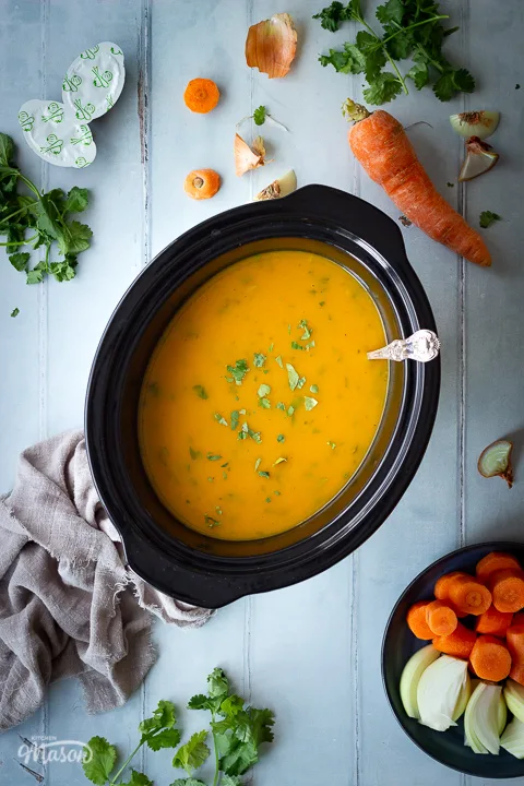 A slow cooker filled with carrot and coriander soup with a ladle inside. There's vegetable peelings scattered around along with a light brown linen napkin, a plate of chopped carrot and onion, some stock pots and coriander. All set on a cool grey wood effect backdrop.