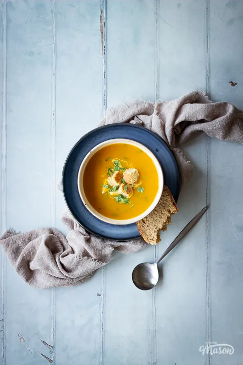 A bowl set on a blue plate over a light brown linen napkin, filled with carrot and coriander soup that's topped with croutons, soured cream and coriander. Set on a cool grey wood effect backdrop, there's also a spoon on the side.