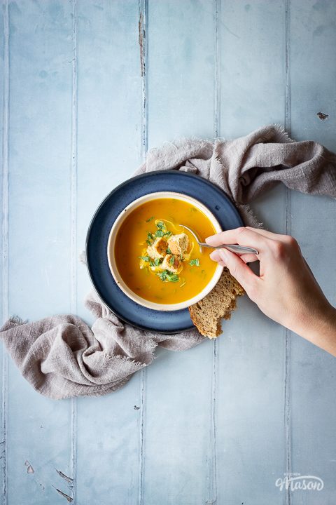 A hand reaching in with a spoon over a bowl set on a blue plate over a light brown linen napkin, filled with carrot and coriander soup that's topped with croutons, soured cream and coriander. Set on a cool grey wood effect backdrop, there's also a spoon on the side and a torn piece of bread.