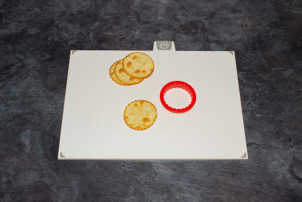 A few crackers on a white chopping board with a fluted circular cutter. Set on a kitchen worktop.