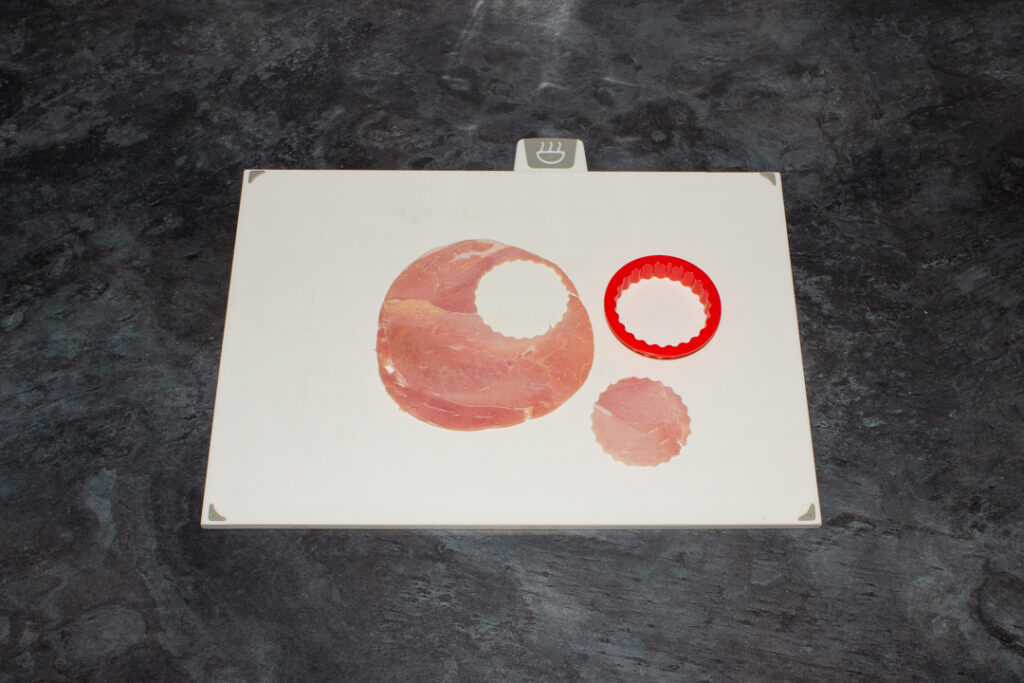 A slice of ham with a fluted circle cut out of it with a fluted circle cookie cutter on a white chopping board. Set on a kitchen worktop.