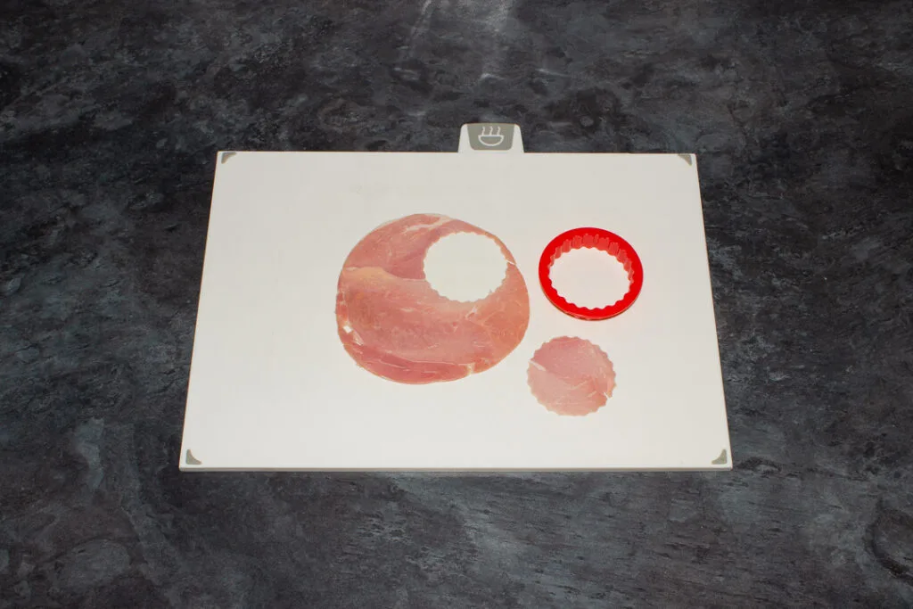 A slice of ham with a fluted circle cut out of it with a fluted circle cookie cutter on a white chopping board. Set on a kitchen worktop.