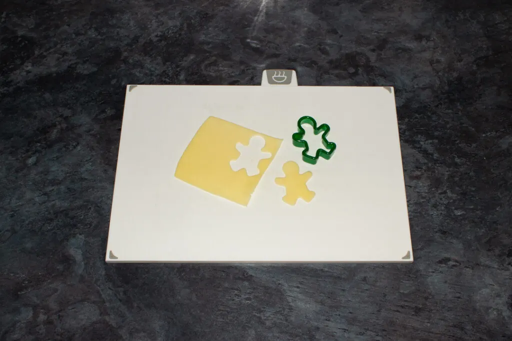 A slice of cheese with a gingerbread man shape cut out of it and a gingerbread man shape cutter on a white chopping board. Set on a kitchen worktop.