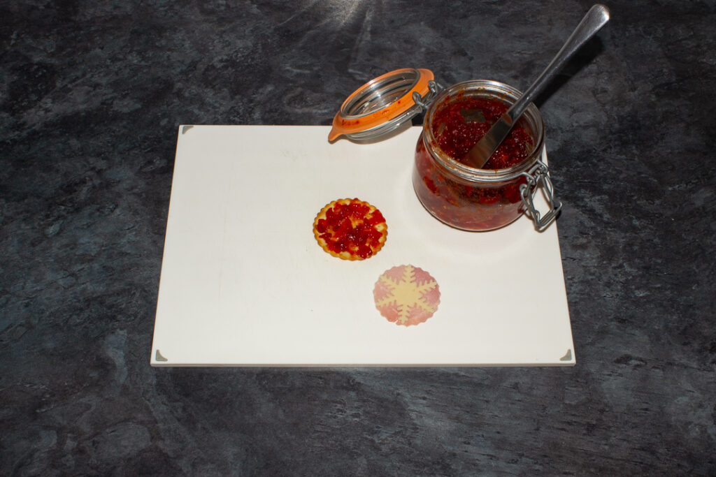 A cracker that's been spread with sweet chilli jam, a jar of sweet chilli jam with a knife in it and a fluted circular piece of ham topped with a cheese snowflake on a white chopping board. Set on a kitchen worktop.