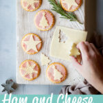 A hand reaching into a white wood board topped with 6 Christmas ham and cheese cracker snacks, a rosemary sprig and a Christmas tree cutter on a slice of cheese. Set on a cool grey wood effect backdrop, there's also more Christmas cutters, some ham and cheese cracker snacks and a light brown linen napkin in the background. A text overlay says "ham and cheese cracker snacks".