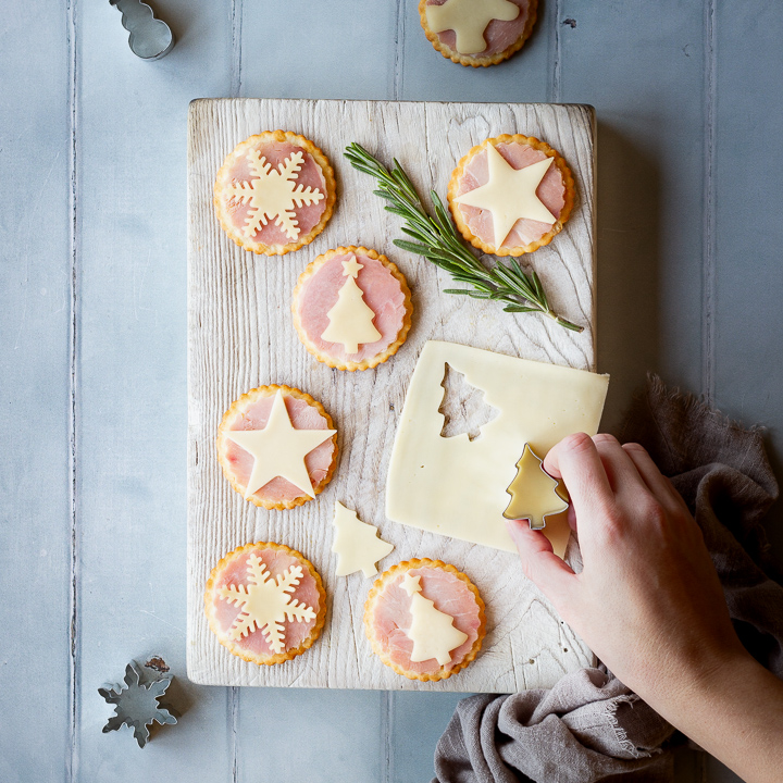 A hand reaching into a white wood board topped with 6 Christmas ham and cheese cracker snacks, a rosemary sprig and a Christmas tree cutter on a slice of cheese. Set on a cool grey wood effect backdrop, there's also more Christmas cutters, some ham and cheese cracker snacks and a light brown linen napkin in the background.