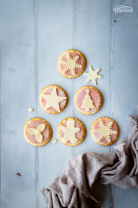6 ham and cheese cracker snacks set on a cool grey wood effect backdrop with a few other cheese shapes scattered around. There's also a light brown linen napkin scrunched up in the corner.