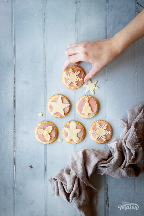 A hand reaching in for 1 of 6 ham and cheese cracker snacks set on a cool grey wood effect backdrop with a few other cheese shapes scattered around. There's also a light brown linen napkin scrunched up in the corner.
