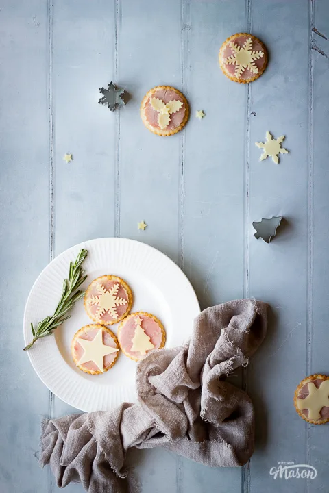 3 ham and cheese cracker snacks set on a white plate with a spring of rosemary in the corner of the shot. Set over a cool grey wood effect backdrop, there's a light brown linen napkin scrunched up at the side of the plate and more ham and cheese cracker snacks, cutters and cheese shapes scattered around in the background.