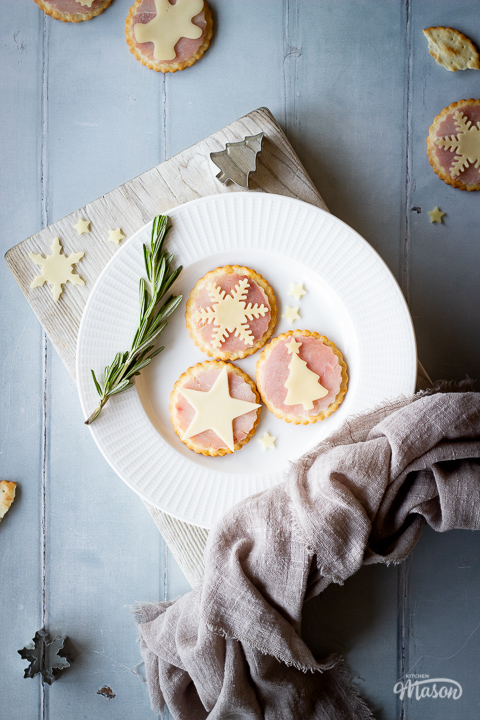3 ham and cheese cracker snacks on a white plate with a sprig of rosemary on the side, set on a white wood chopping board. There are cheese shapes, a Christmas tree cutter and a light brown linen napkin on the edges of the board. Set over a cool grey wood effect backdrop, there are more ham and cheese cracker snacks and cutters scattered in the background.