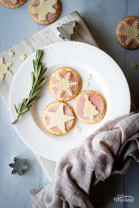 Close up of 3 ham and cheese cracker snacks on a white plate with a sprig of rosemary on the side, set on a white wood chopping board. There are cheese shapes, a Christmas tree cutter and a light brown linen napkin on the edges of the board. Set over a cool grey wood effect backdrop, there are more ham and cheese cracker snacks and cutters scattered in the background.