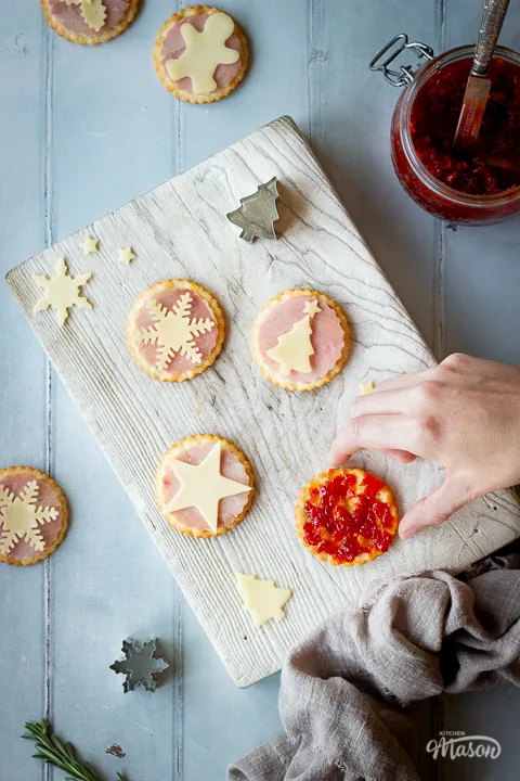 A hand reaching over a white wood chopping board with ham and cheese cracker snacks, cheese shapes, a Christmas tree cutter and a cracker spread with sweet chilli jam on top. Set on a cool grey wood effect backdrop, there's also a light brown linen napkin, a jar of sweet chilli jam with a knife in it and more cutters scattered in the background.