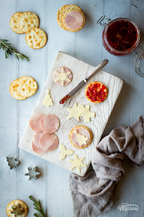 Circles of ham, Christmas cheese shapes, a cracker with sweet chilli jam on it and a knife on a white wood chopping board set on a cool grey wood effect backdrop. There are also crackers, rosemary sprigs, cutters, a jar of sweet chilli jam and a light brown linen napkin in the background.