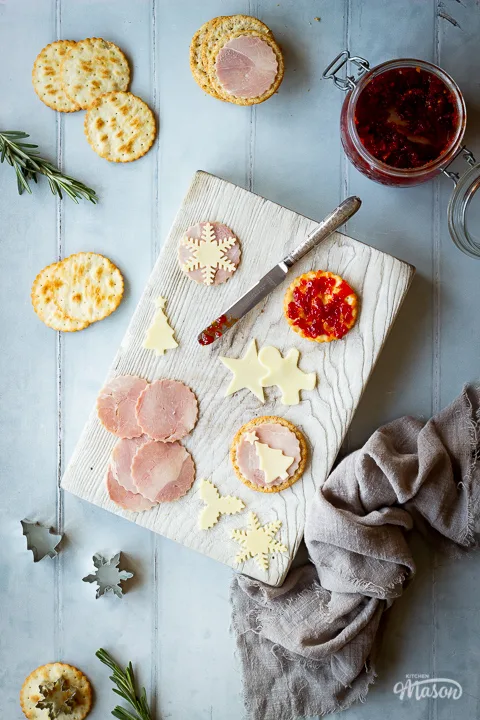 Circles of ham, Christmas cheese shapes, a cracker with sweet chilli jam on it and a knife on a white wood chopping board set on a cool grey wood effect backdrop. There are also crackers, rosemary sprigs, cutters, a jar of sweet chilli jam and a light brown linen napkin in the background.