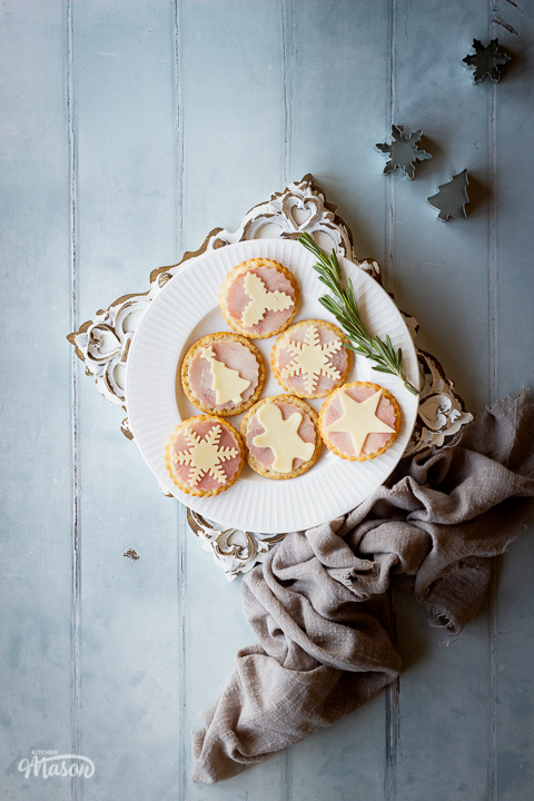 6 Christmas ham and cheese cracker snacks on a white plate with a spring of rosemary set on an ornate brushed white wood board. All set on a cool grey wood effect backdrop, there's also a light brown linen napkin and some cutters in the background.