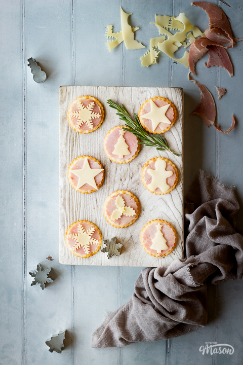 A white wood board with 8 Christmas ham and cheese cracker snacks, a rosemary sprig and a snowflake cutter on top. Set on a cool grey wood effect backdrop, there's also more Christmas cutters, some ham and cheese scraps and a light brown linen napkin in the background.