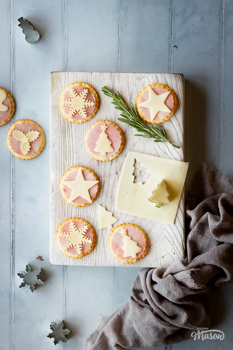 A white wood board topped with 6 Christmas ham and cheese cracker snacks, a rosemary sprig and a Christmas tree cutter on a slice of cheese. Set on a cool grey wood effect backdrop, there's also more Christmas cutters, some ham and cheese cracker snacks and a light brown linen napkin in the background.