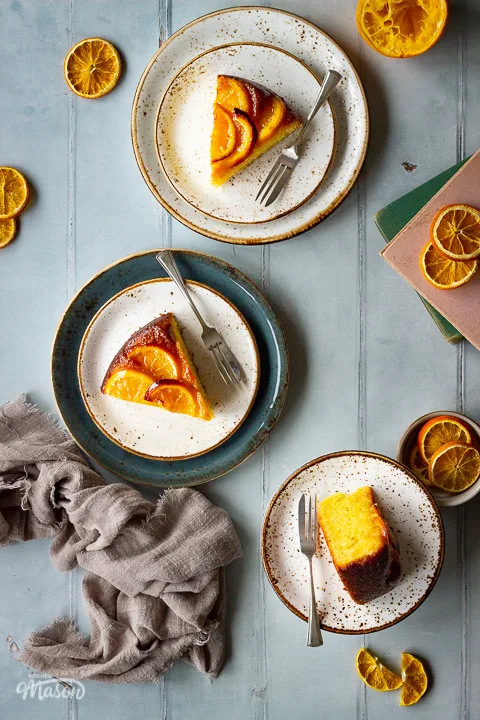 A flat lay view of 3 plates, each with a slice of sticky orange cake and a fork on them, set over a cool grey wood effect backdrop. There are also dried orange slices, two stacked books, half a squeezed orange and a light brown linen napkin in the background.