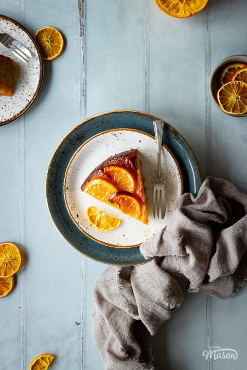 Flay lay view of a slice of sticky orange cake on two stacked plates with a fork. Set over a cool grey wood effect backdrop, there are also dried orange slices, another plate with cake, a small pot filled with orange slices and a light brown linen napkin in the background.