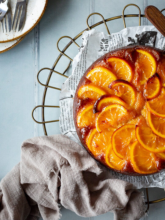 A whole sticky orange cake on a round cooling rack over a cool grey wood effect backdrop. There are also dried orange slices, white rustic plates with dessert forks, two stacked books and a light brown linen napkin in the background.