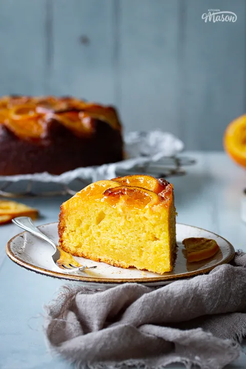 A front view of a slice of sticky orange cake on a plate with a fork positioned to the centre of the shot. In the background there is the remaining sticky orange cake on a round cooling rack, half a squeezed orange, some dried orange slices and a light brown linen napkin. All set over a cool grey wood effect backdrop.