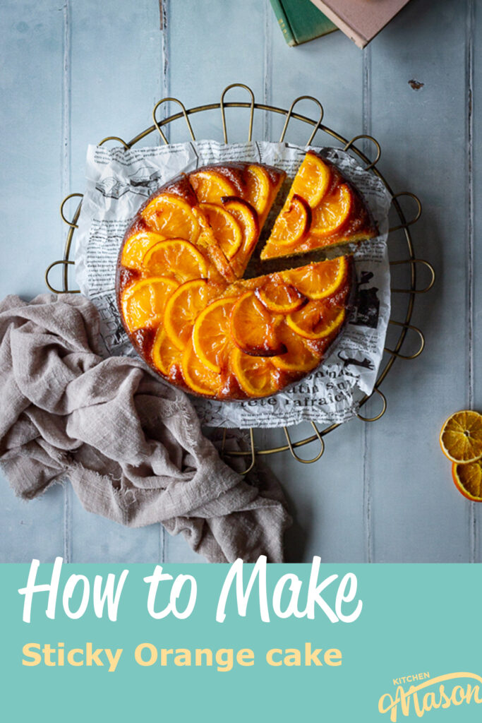 A flat lay of a sticky orange cake with a slice cut out of it on a round cooling rack over a cool grey wood effect backdrop. There are also dried orange slices, two stacked books and a light brown linen napkin in the background. A text overlay says "How to make sticky orange cake".