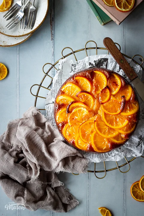 A whole sticky orange cake on a round cooling rack positioned to the right side over a cool grey wood effect backdrop. There are also dried orange slices, white rustic plates with dessert forks, two stacked books and a light brown linen napkin in the background.