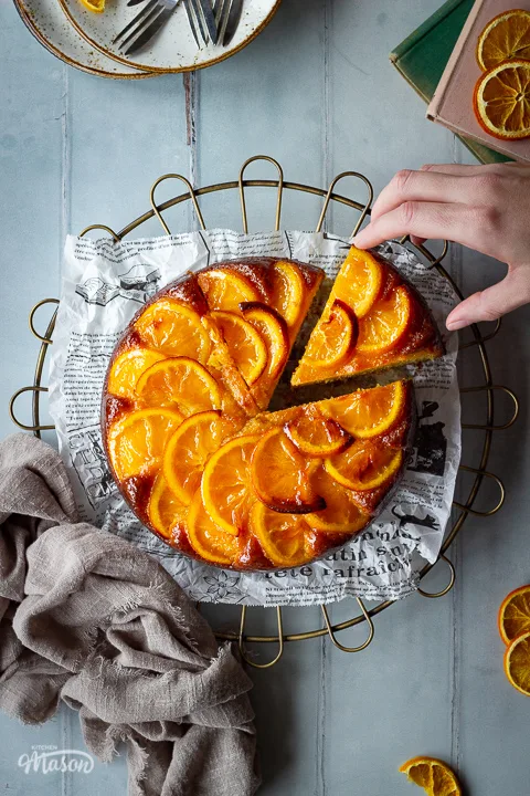 A sticky orange cake with a slice cut out of it and a hand reaching in on a round cooling rack over a cool grey wood effect backdrop. There are also dried orange slices, white rustic plates with dessert forks, two stacked books and a light brown linen napkin in the background.