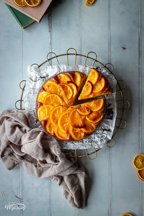 A distanced flat lay of a sticky orange cake with a slice cut out of it on a round cooling rack over a cool grey wood effect backdrop. There are also dried orange slices, two stacked books and a light brown linen napkin in the background.
